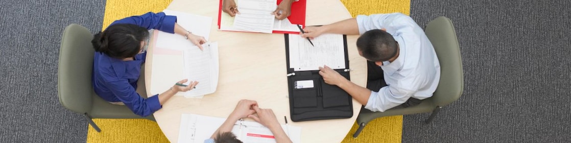  Cuatro empresarios sentados alrededor de una mesa redonda leyendo documentos, alfombra gris y amarilla