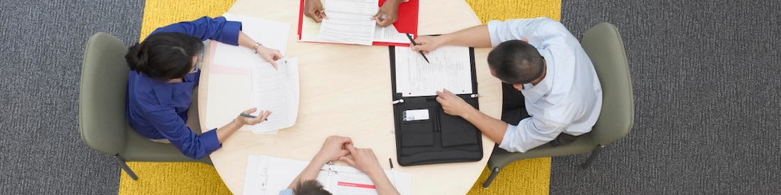 Cuatro empresarios sentados alrededor de una mesa redonda leyendo documentos, alfombra gris y amarilla