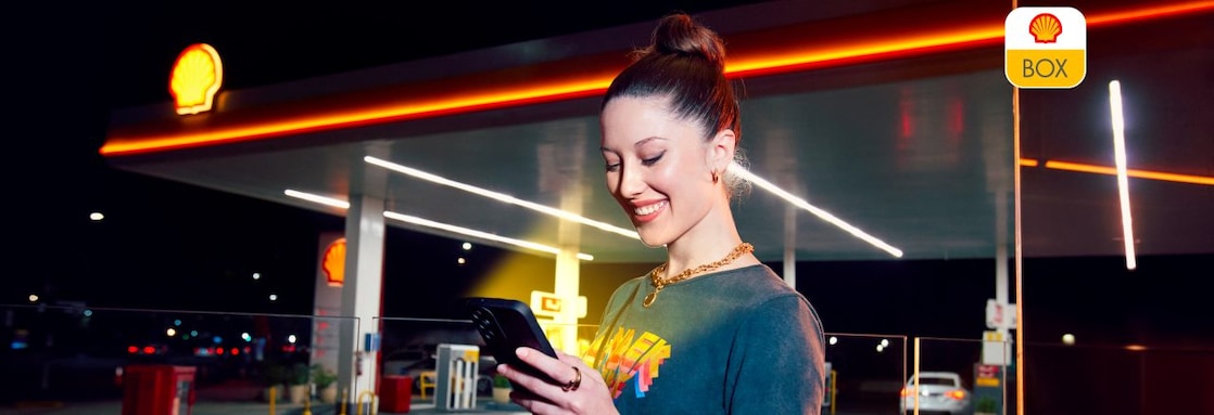 Mujer con el celular y estación de servicio Shell de fondo.
