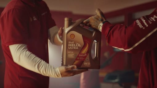 Dos personas con uniformes de escudería se pasan un envase de aceite Shell Helix Ultra. 