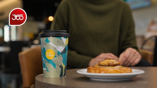 Vaso de café y roll de canela sobre una mesa en Shell Select. Logo de Clarín 365 en la esquina superior izquierda.