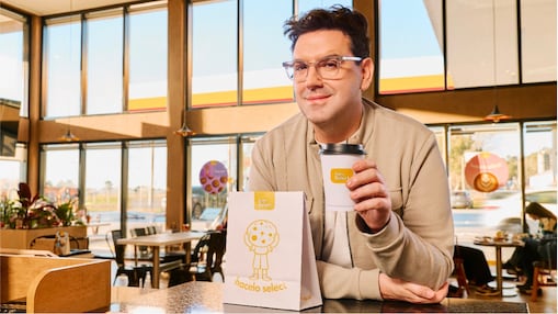 Damián Betular sosteniendo un vaso de café en el mostrador de Shell Select junto a una bolsa de pastelería.
