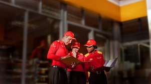 Tres empleados (un hombre y dos mujeres) con uniforme de Shell -camisa roja y gorra- examinan una carpeta que sostiene el hombre. Una de las mujeres también lleva en la mano un maletín con documentos.