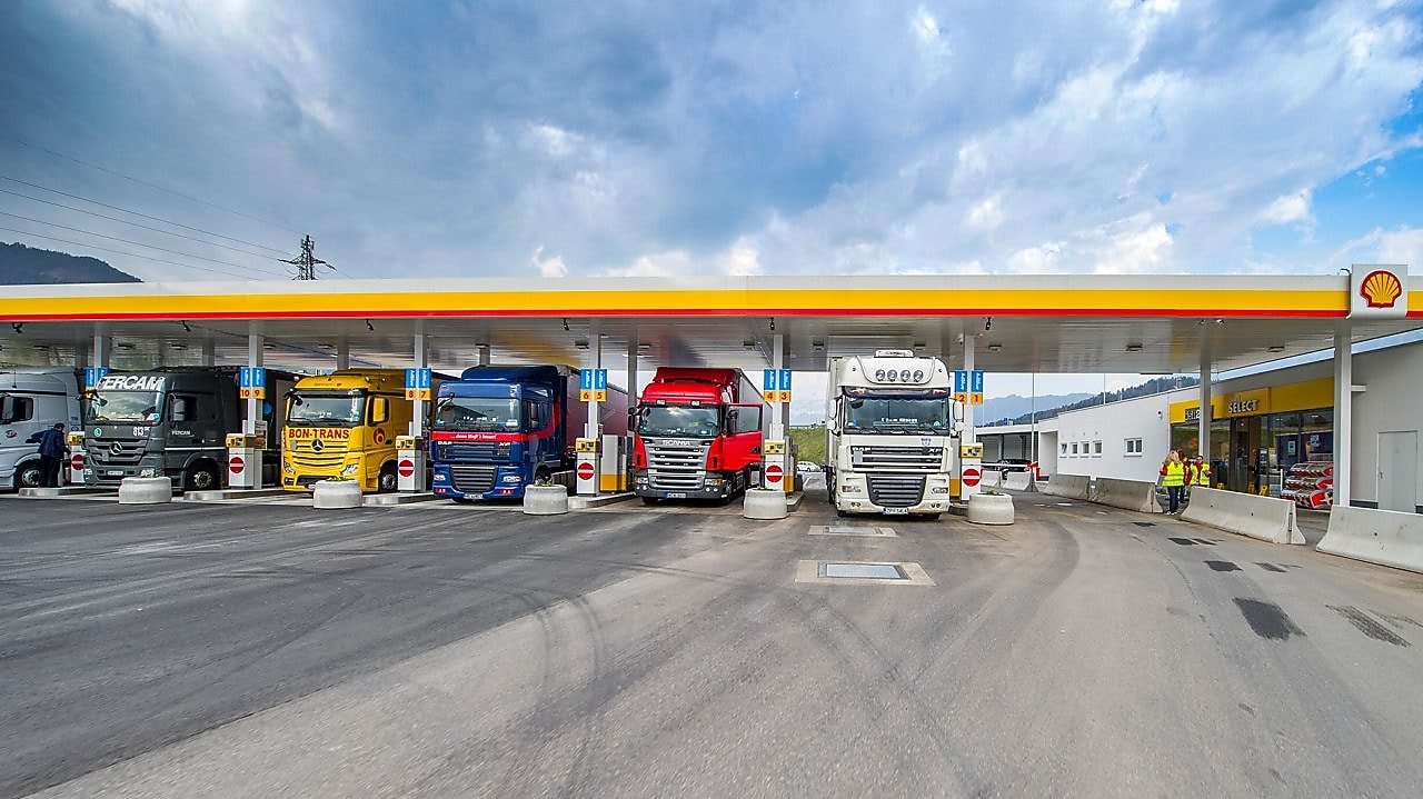 Gasolinera Shell con seis camiones aparcados. De izquierda a derecha: un camión blanco, verde oscuro, amarillo, azul oscuro, rojo y otro camión blanco.